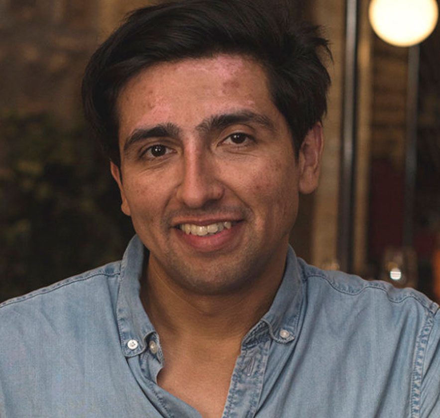 A man with dark hair and a denim shirt smiles at the camera in an indoor setting with dim lighting.