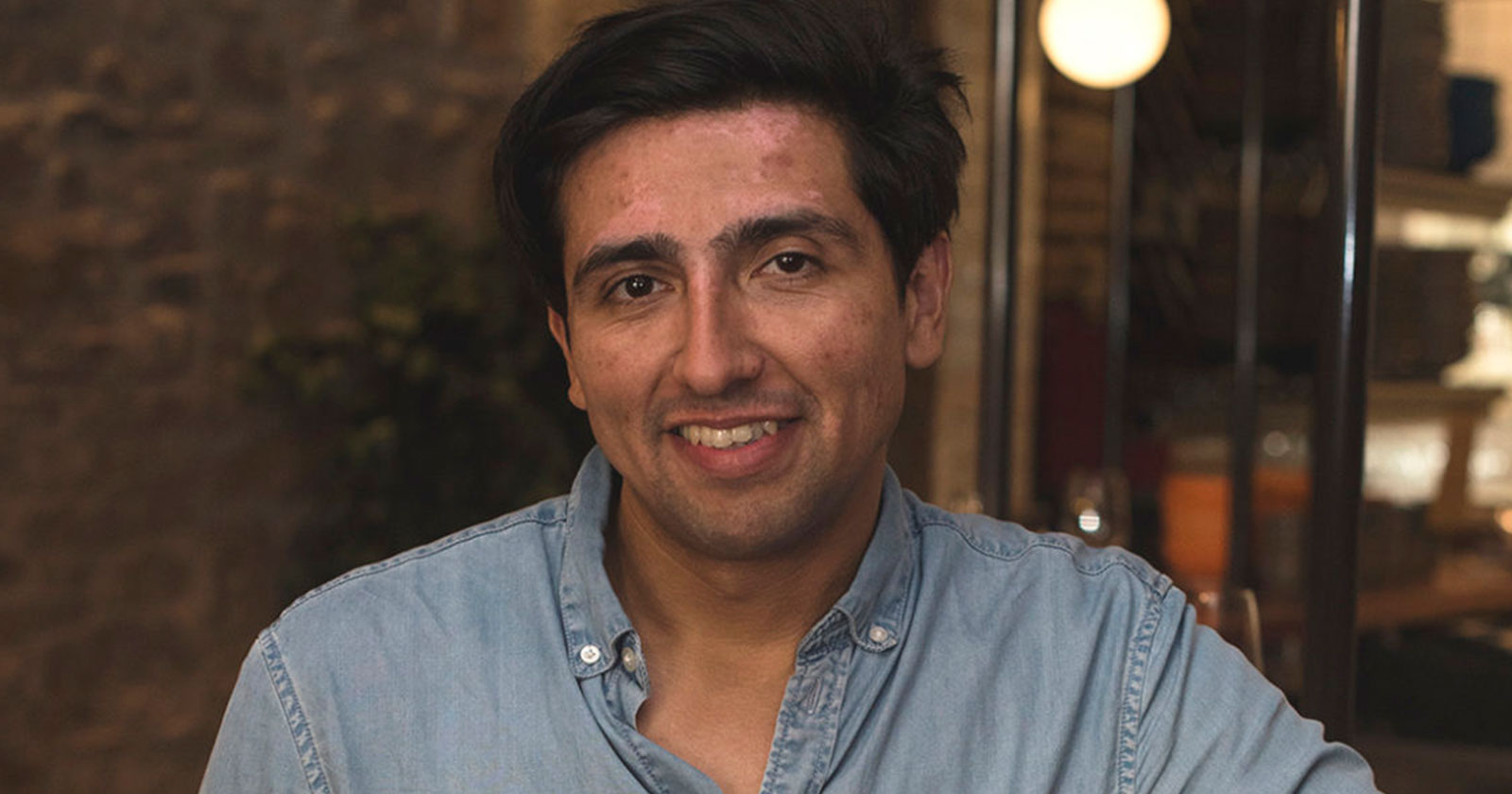 A man with dark hair and a denim shirt smiles at the camera in an indoor setting with dim lighting.