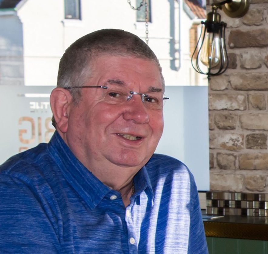 A man wearing glasses and a blue shirt sits at a table indoors with a glass of Heineken beer in front of him. There is a window and a brick wall in the background.