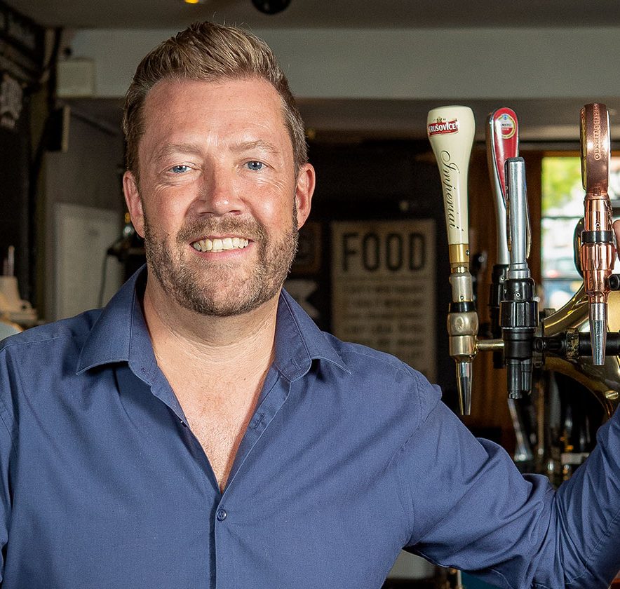 Man in blue shirt smiling and standing next to beer taps in a bar.