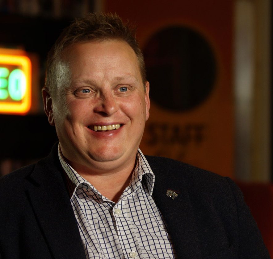 A person in a suit is sitting and smiling in front of a blurred background with a neon "VIDEO" sign.