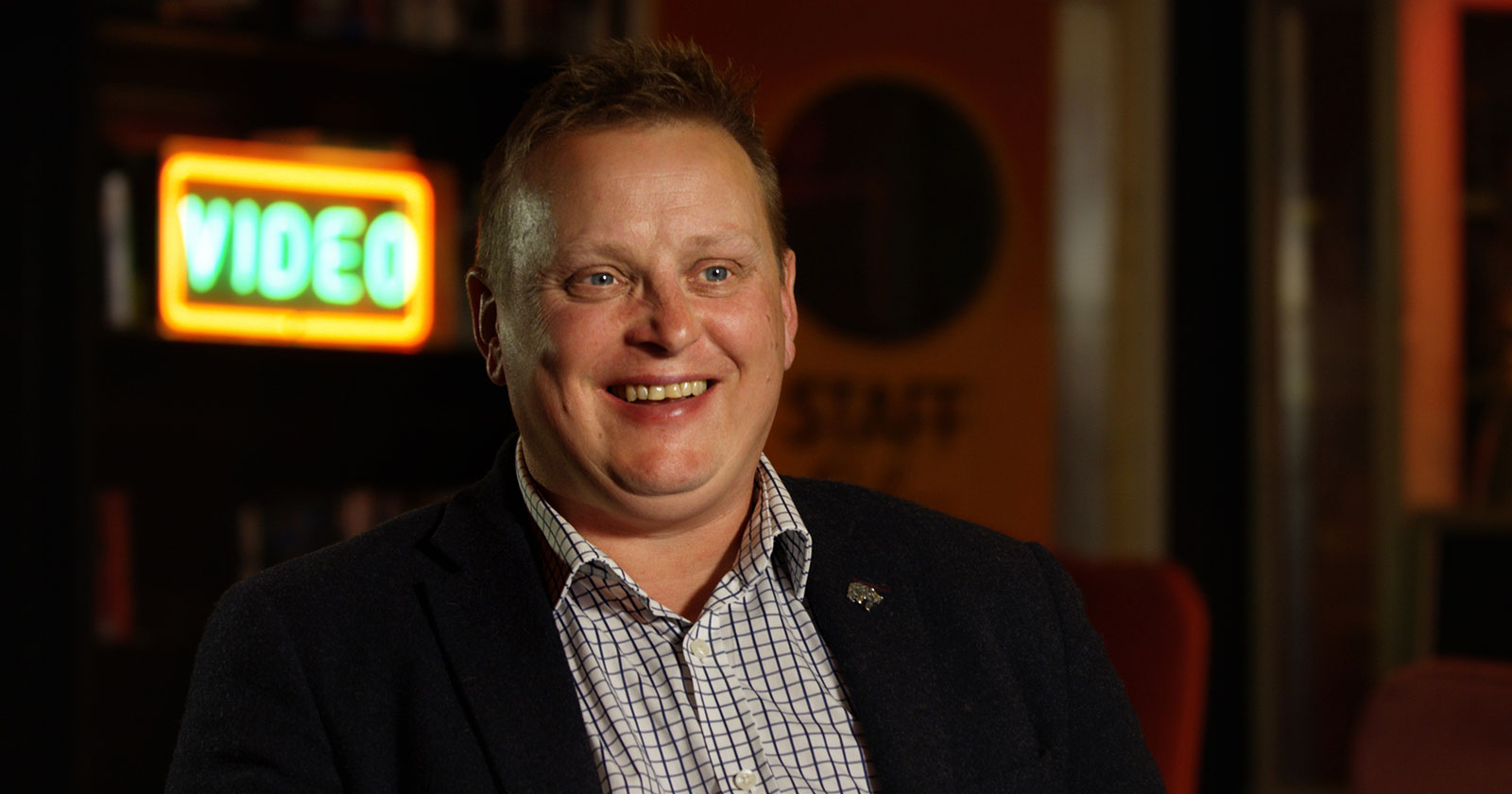 A person in a suit is sitting and smiling in front of a blurred background with a neon "VIDEO" sign.