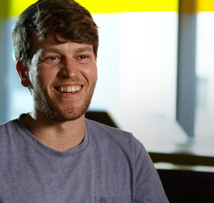 A man with a beard, wearing a blue T-shirt, is smiling while seated in a room with large windows featuring a yellow trim.