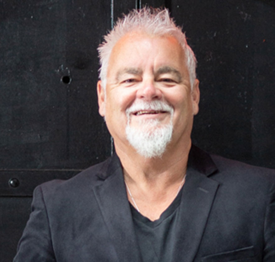 A person with short, spiked, white hair and a white beard, wearing a black blazer over a black shirt.