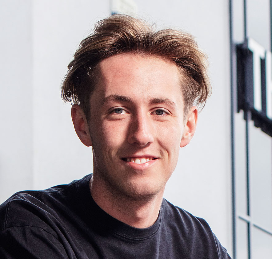 A young man with light brown hair and a black shirt smiles while looking at the camera.