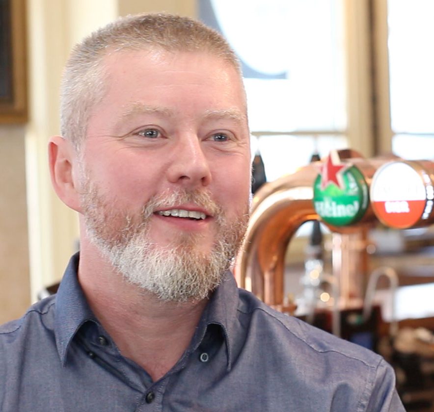 A man with a gray beard and short hair smiles while sitting in a bar. Various beer taps are visible in the background.