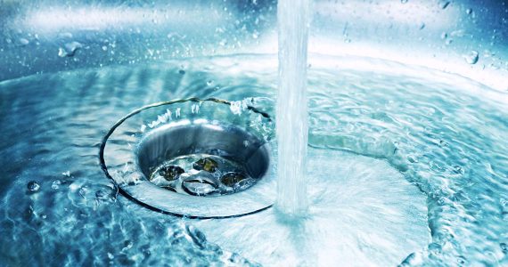 Water flowing from a faucet into a stainless steel sink, creating ripples and splashing around the drain hole.