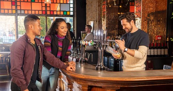 Two people stand at a bar while a bartender pours a drink from a tap. The bar interior features colorful stained glass and various bottles.