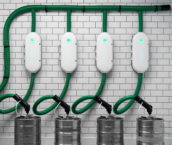 Four wall-mounted devices connected by green hoses to four metal kegs, set against a white tiled background.