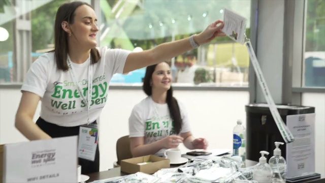 Two women at a registration desk in a well-lit room. They are wearing white t-shirts with green text and handing out items, including name tags. Various supplies are on the table.